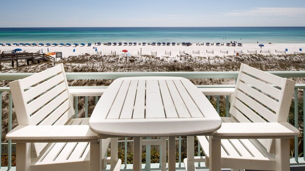 A beach view from a deck with white chairs and a table, overlooking a sandy beach with umbrellas and people. - PI 307: Sunkissed & Vibing. Absolutely beautiful, Gulf front, WiFi (Fort Walton Beach)