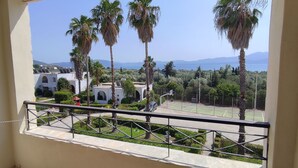 A balcony with a view of a tennis court, palm trees, and a distant mountain range. - Silver Bay Hotel & Bungalows (Lesvos)
