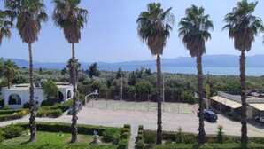 A tennis court surrounded by palm trees and a parking area with cars. - Silver Bay Hotel & Bungalows (Lesvos)