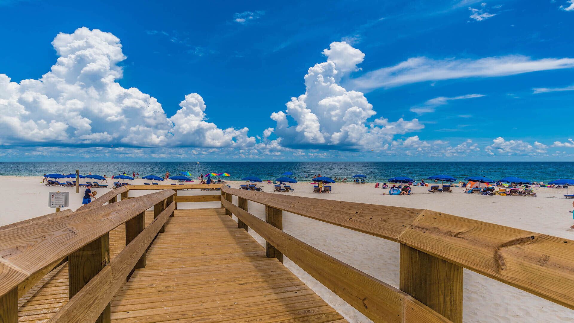 A wooden boardwalk leading to a sandy beach with blue umbrellas and people relaxing.