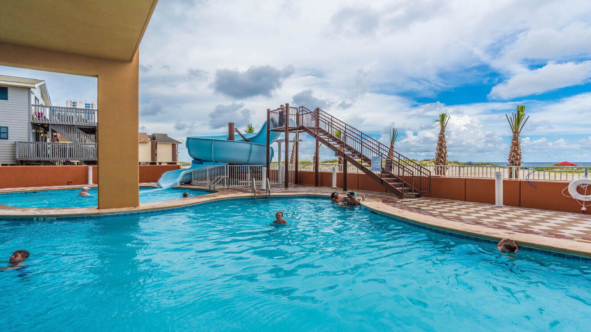 A swimming pool with a water slide, palm trees, and a beach view.