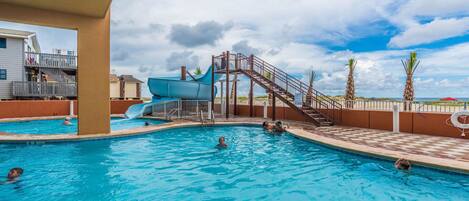 A swimming pool with a water slide, palm trees, and a beach view.