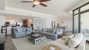 A spacious living area with a large gray sectional sofa, a coffee table, and a TV. There is a ceiling fan, a dining area with a chandelier, and a kitchen in the background.