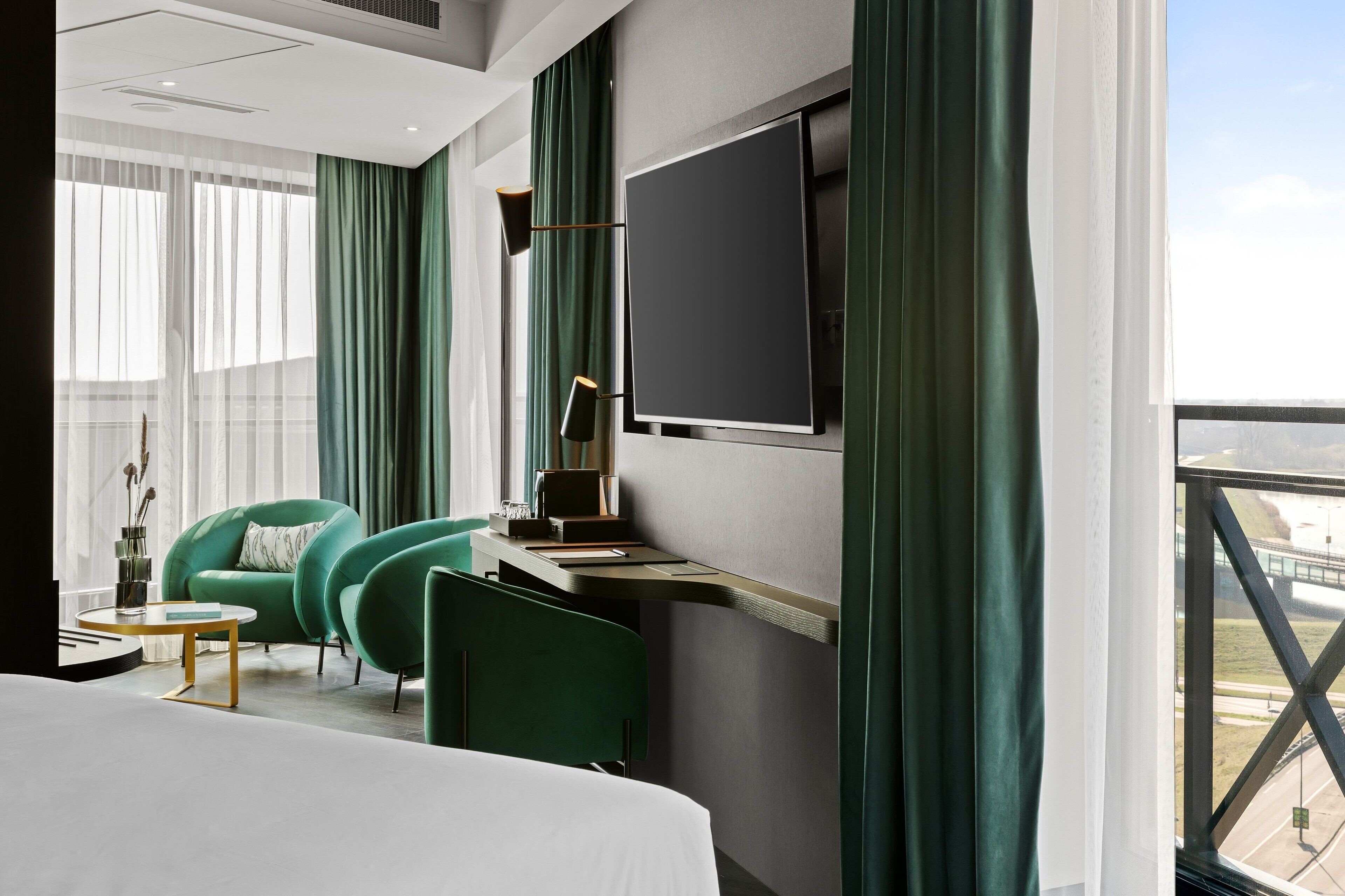 A modern hotel room with a large bed, two green armchairs, a small table, a desk with a flat-screen TV, and a view of the cityscape through a large window.