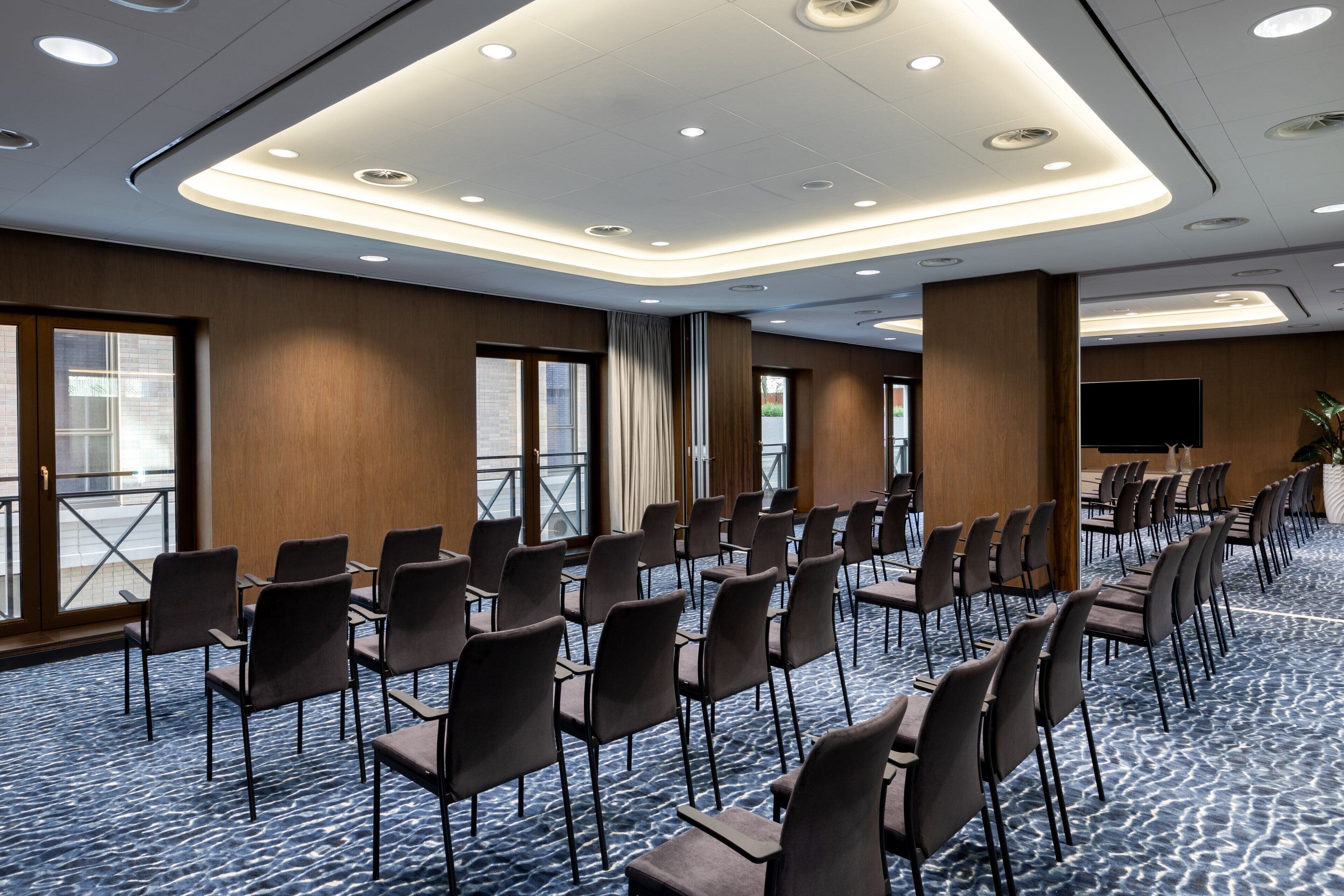Un salón de conferencias con filas de sillas, un ventanal grande y un televisor de pantalla plana.