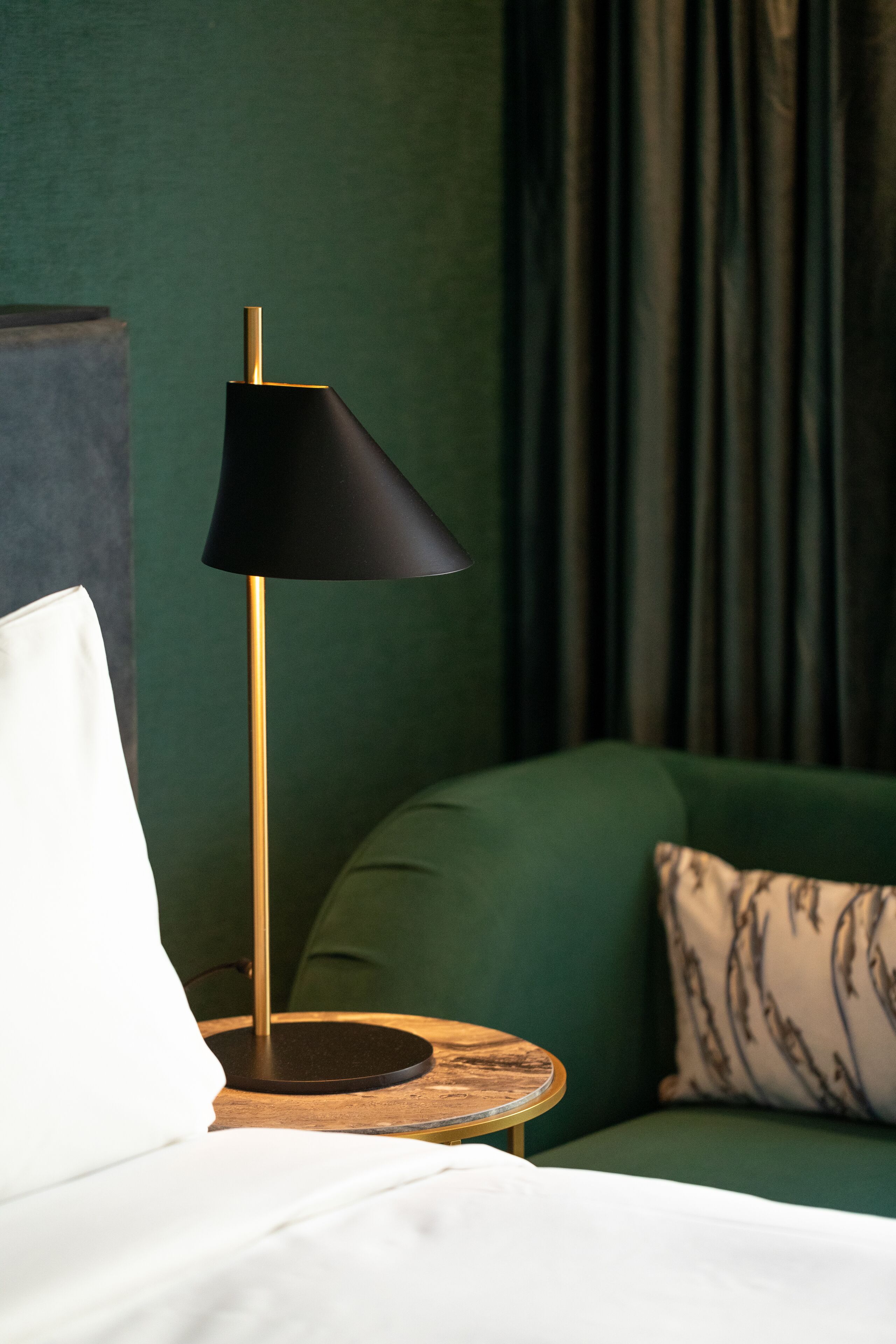 A neatly made bed with a white pillow, a black floor lamp with a gold base, and a green sofa with a patterned pillow.