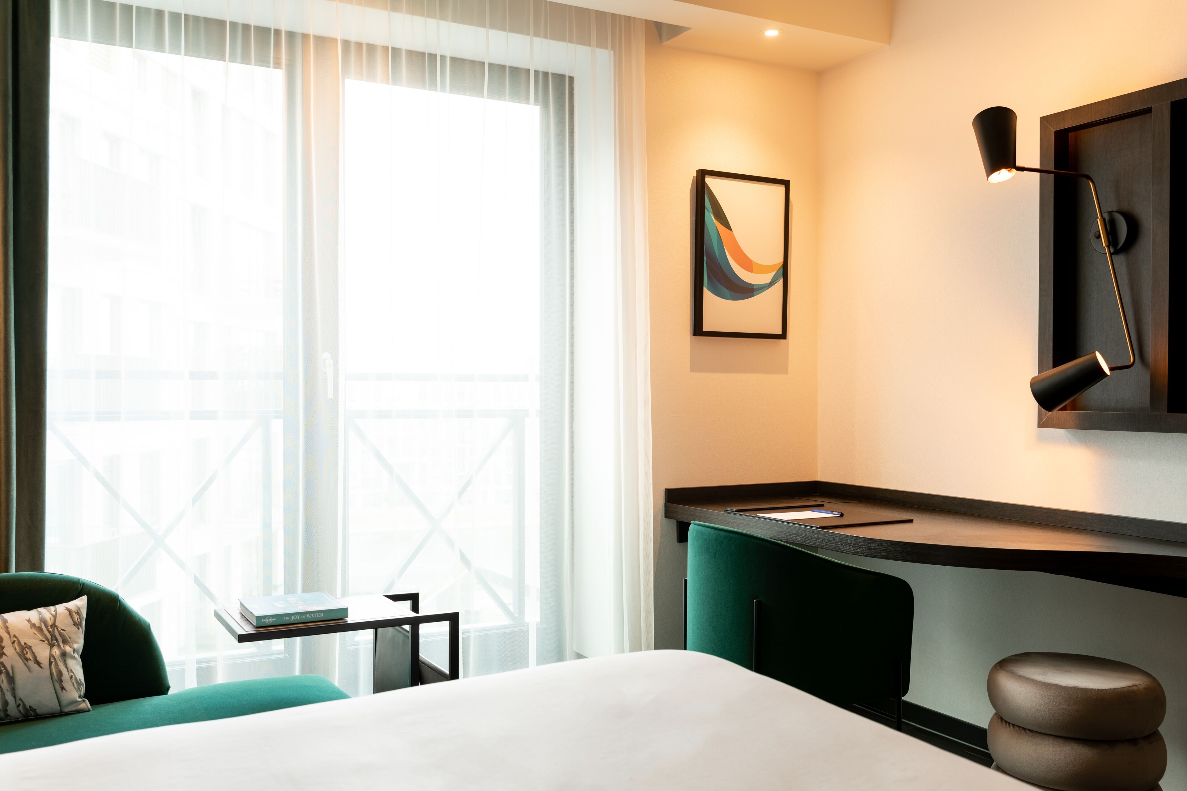 Habitación de hotel con cama, escritorio, silla, ventana con cortinas y lámpara de pared.