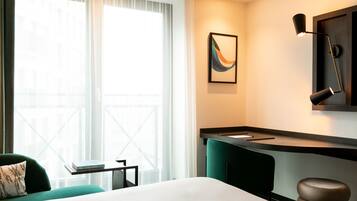 Habitación de hotel con cama, escritorio, silla, ventana con cortinas y lámpara de pared.