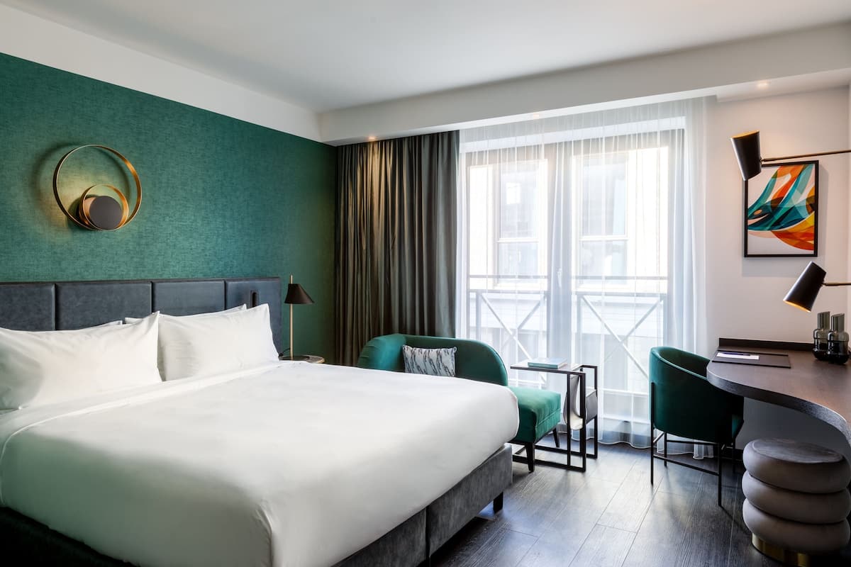 A modern hotel room with a large bed, a desk, and a view of the city through the window.