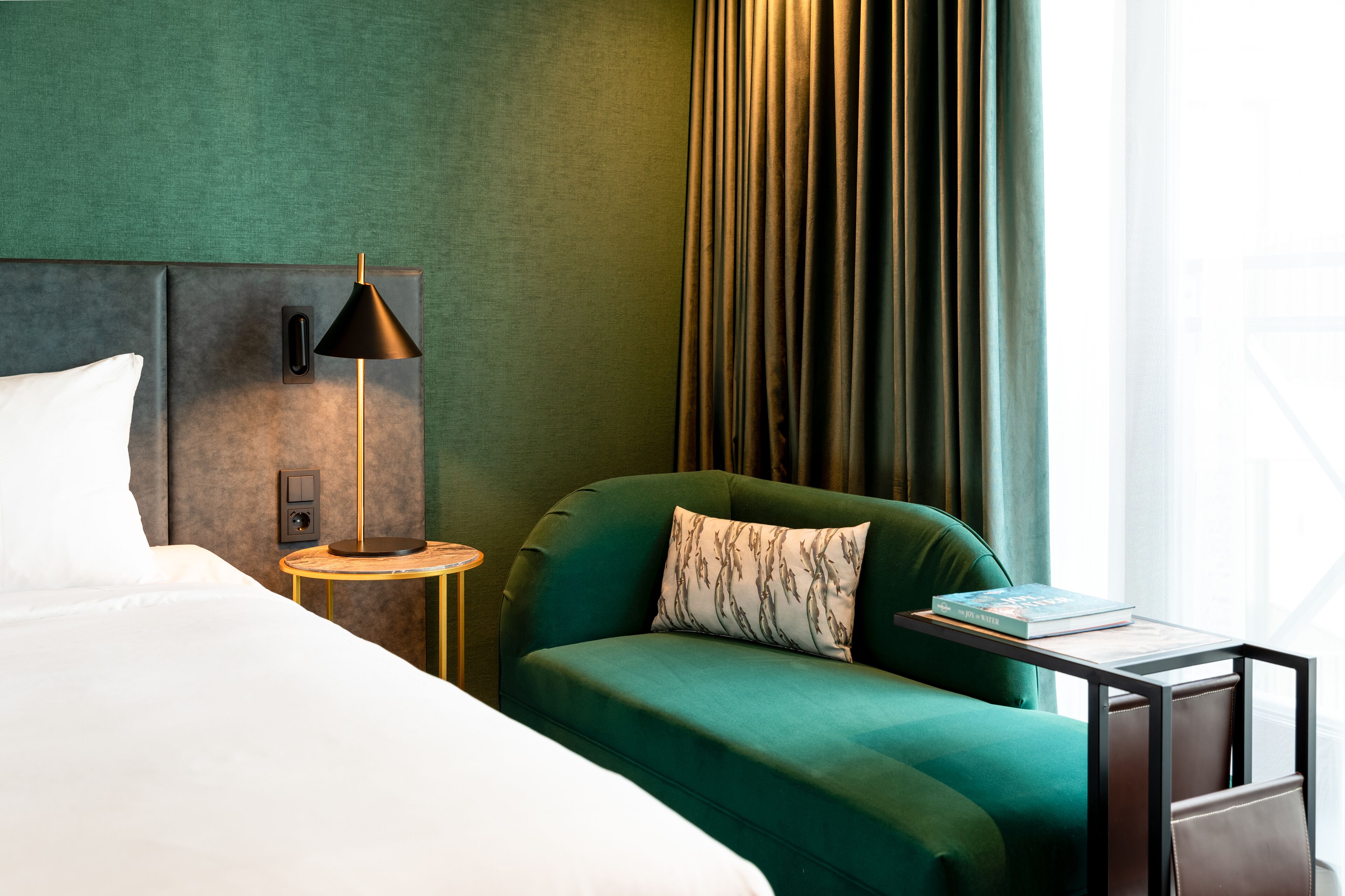 Ett hotellrum med en säng, en grön fåtölj, ett litet runt bord med en lampa och utsikt över omgivningarna genom ett fönster.