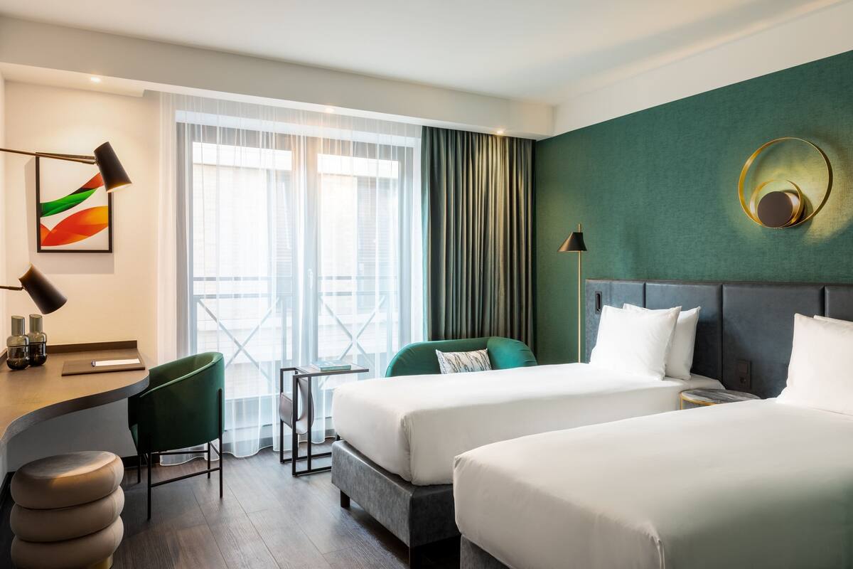 A hotel room with two beds, a desk, a chair, and a view of the city.