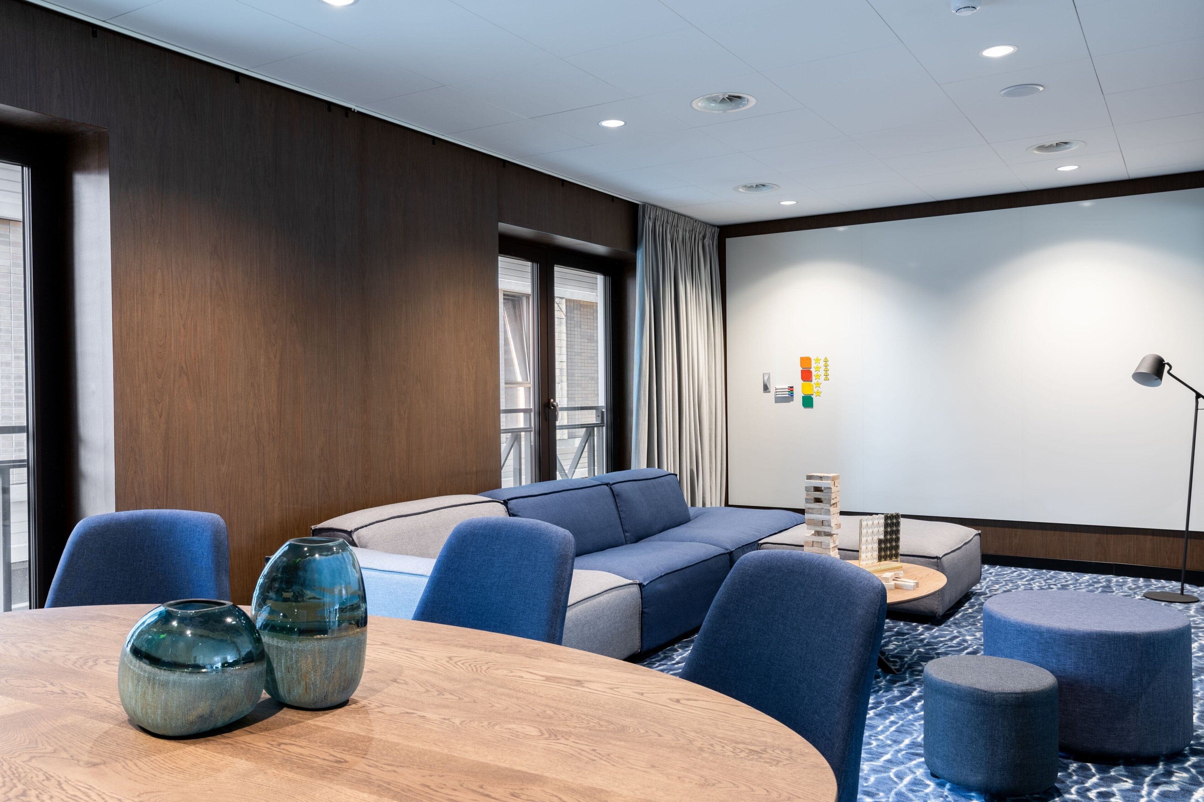 Una habitación de hotel moderna con una pared de acento de madera, un amplio sofá modular, una mesa redonda con dos jarrones y una pantalla de proyector en la pared.