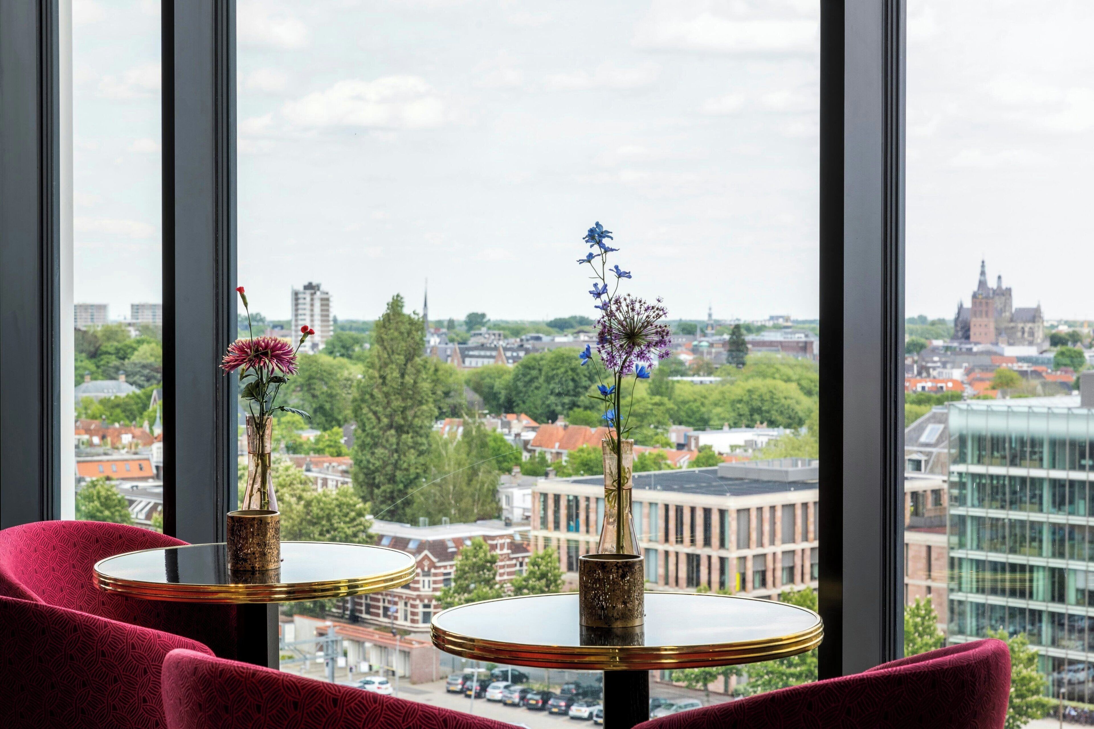 Une pièce dotée de deux tables rondes, chacune ornée d’un vase de fleurs, et offrant une vue sur le paysage urbain grâce à de grandes fenêtres.