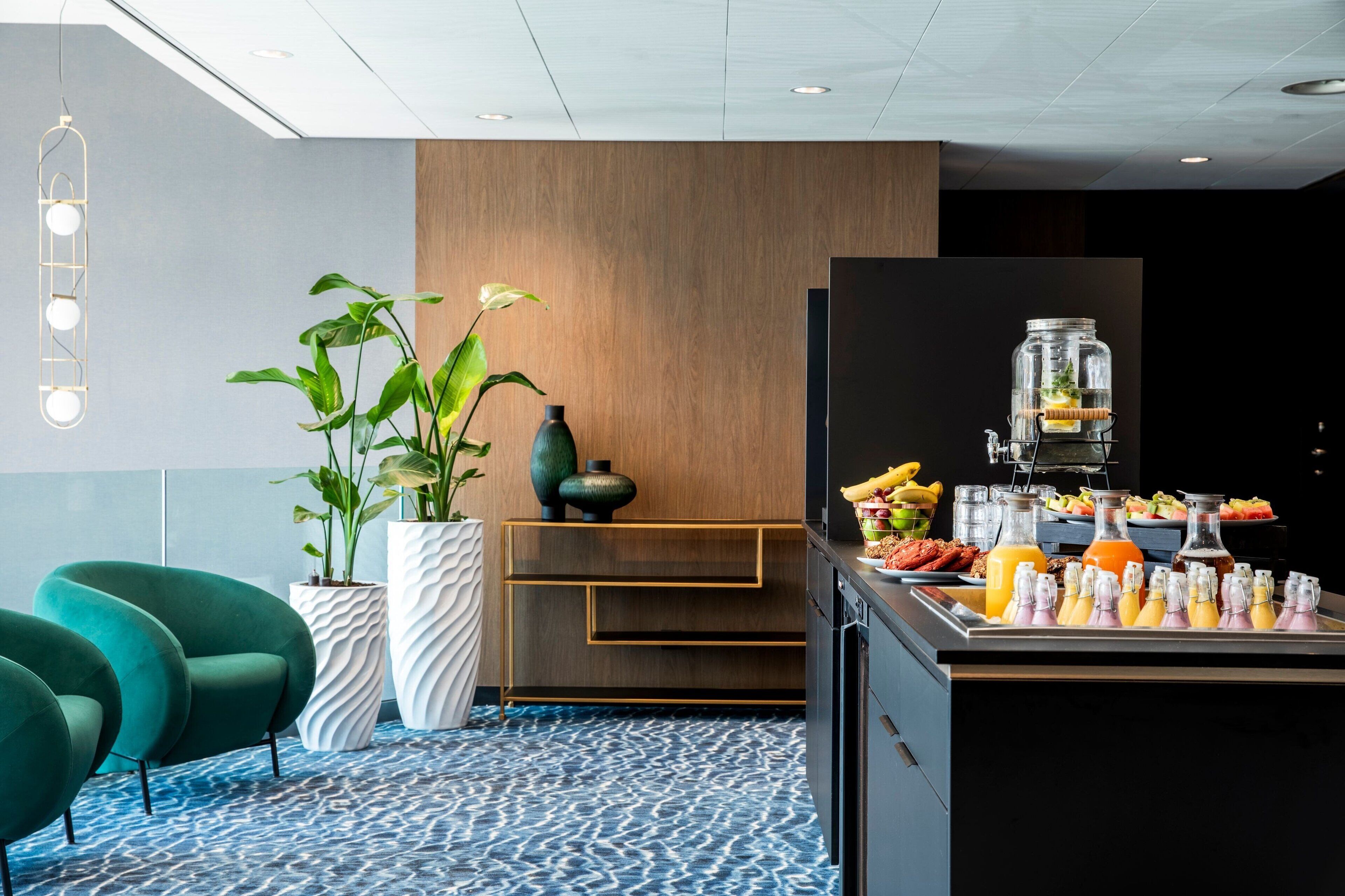 A modern hotel lobby with teal armchairs, a large potted plant, a wooden accent wall, and a breakfast bar with various beverages and food items.
