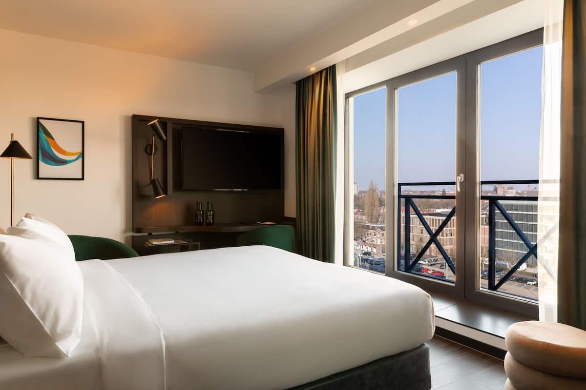 A hotel room with a bed, a flat-screen TV, a desk with a chair, and a large window offering a city view.