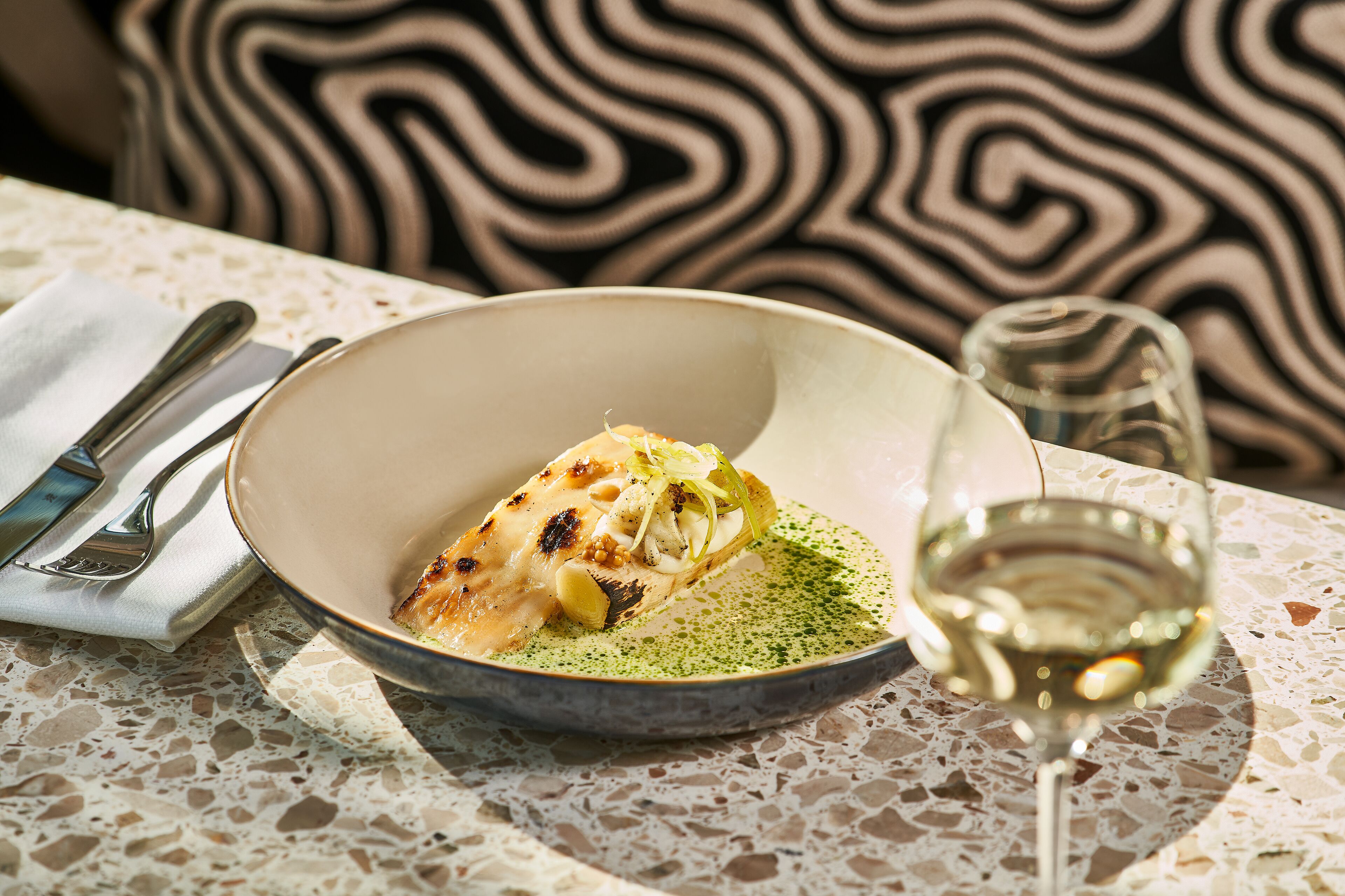A plate of food with a glass of white wine on a table with a patterned tablecloth.