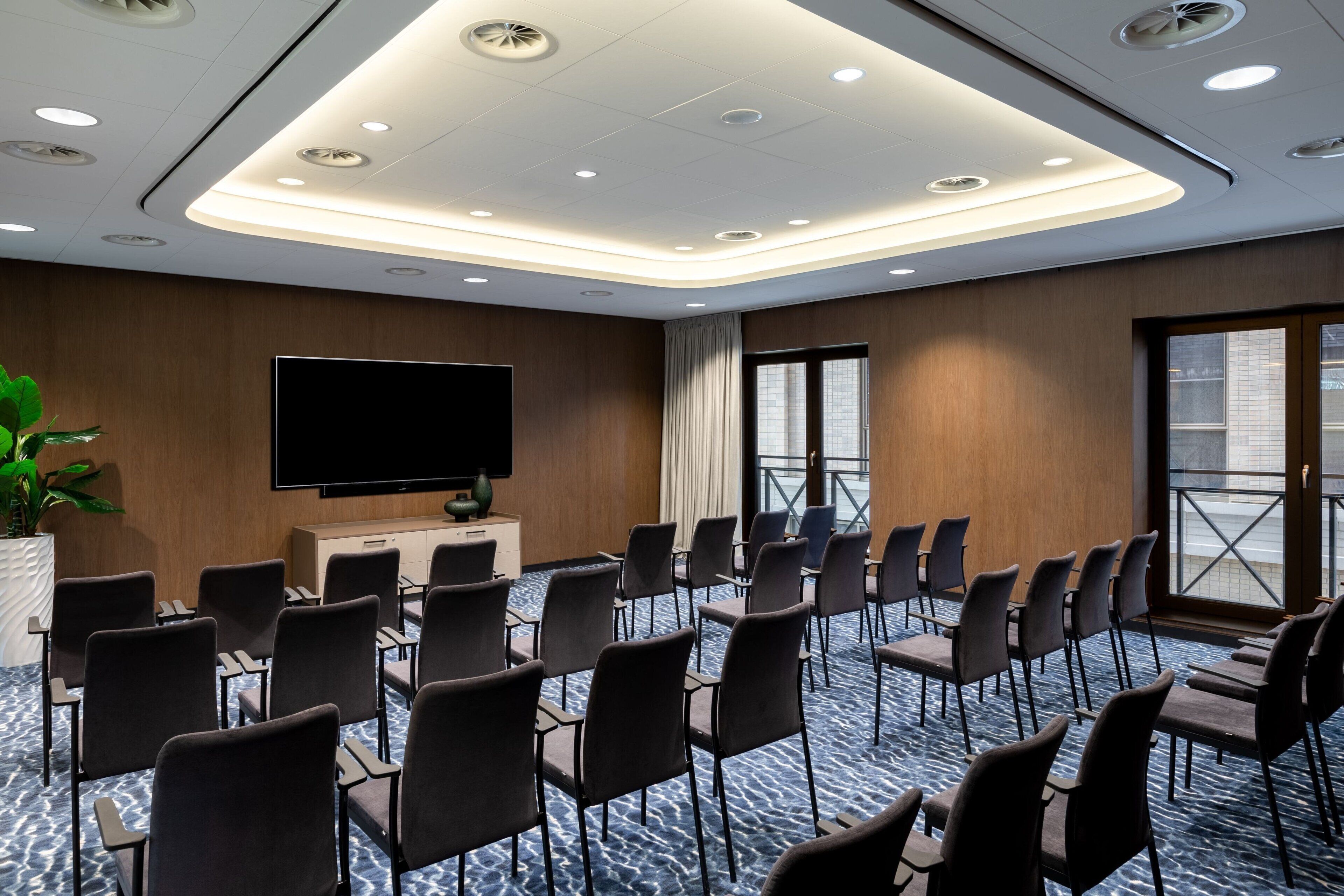 A conference room with a flat-screen TV, rows of chairs, a potted plant, and a window with a view.