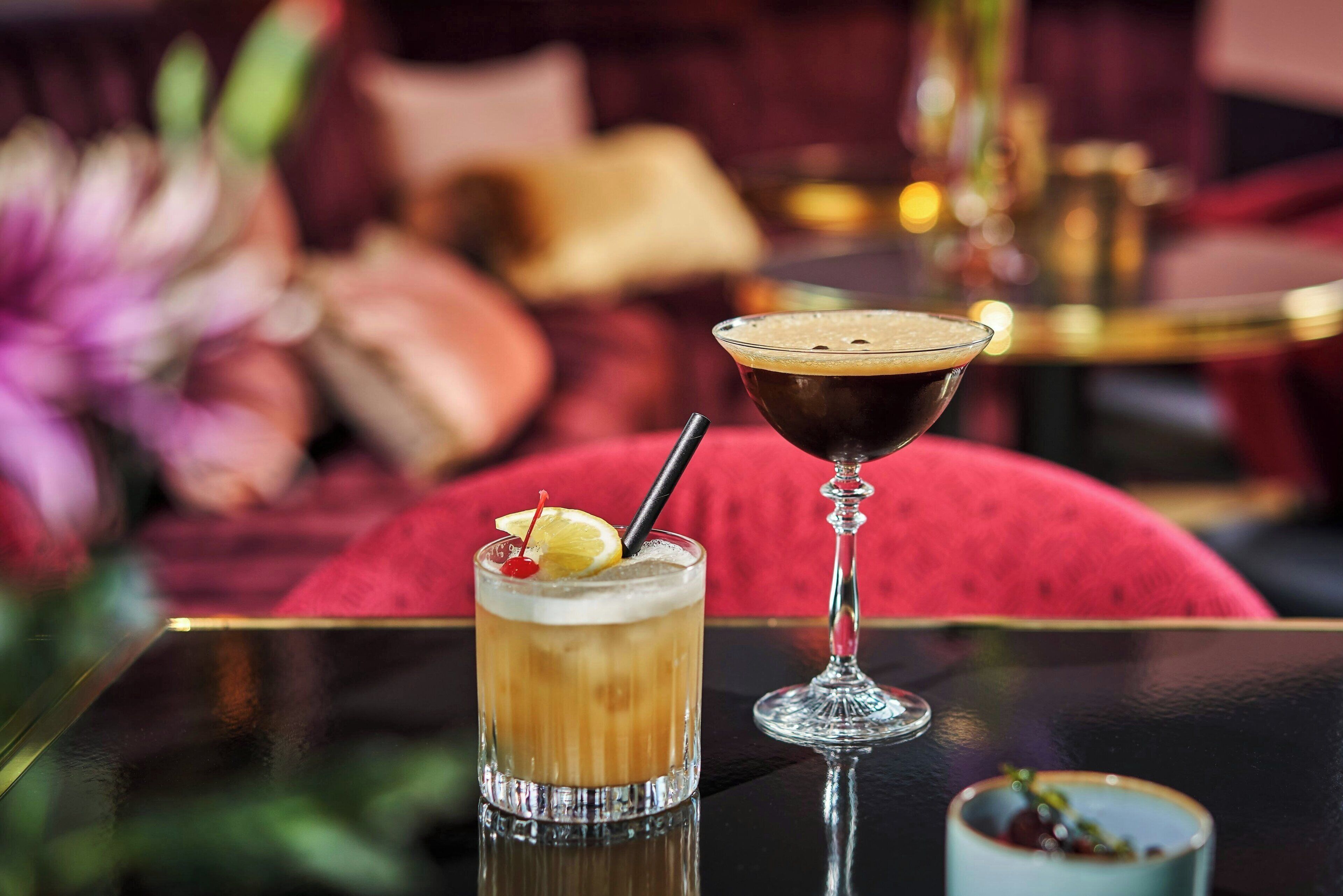 Un verre à martini et un grand verre à cocktail sur une table réfléchissante, avec un arrière-plan flou comprenant un canapé rouge et un vase rempli de fleurs.