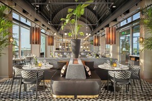 A modern restaurant with a central bar area, multiple seating options, and large windows providing views of the city.