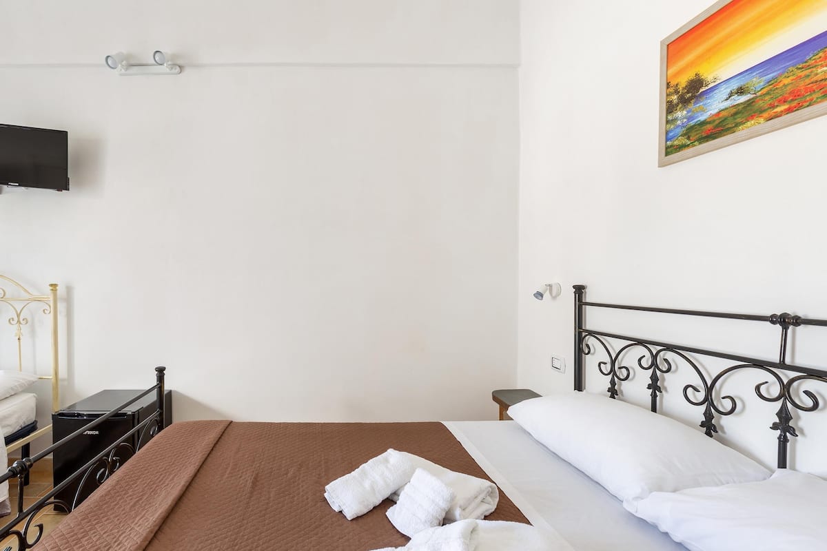 Una cama bien hecha con cubrecama marrón y almohadas blancas, una mesita de noche negra con una lámpara, un televisor montado en la pared y un cuadro de un paisaje.