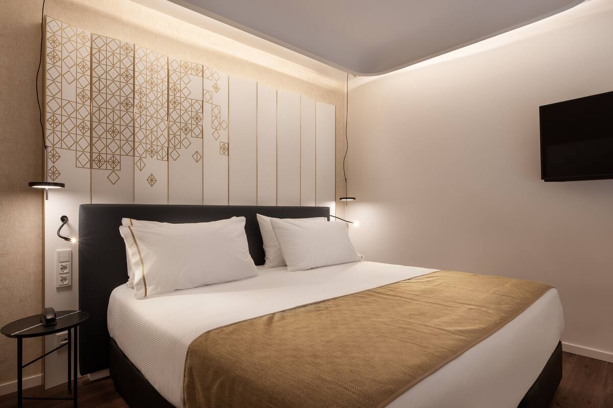 Ein modernes Hotelzimmer mit einem großen Bett, einem Nachttisch mit Lampe, einem Wand-Fernseher und einer dekorativen Wandverkleidung mit geometrischem Muster.