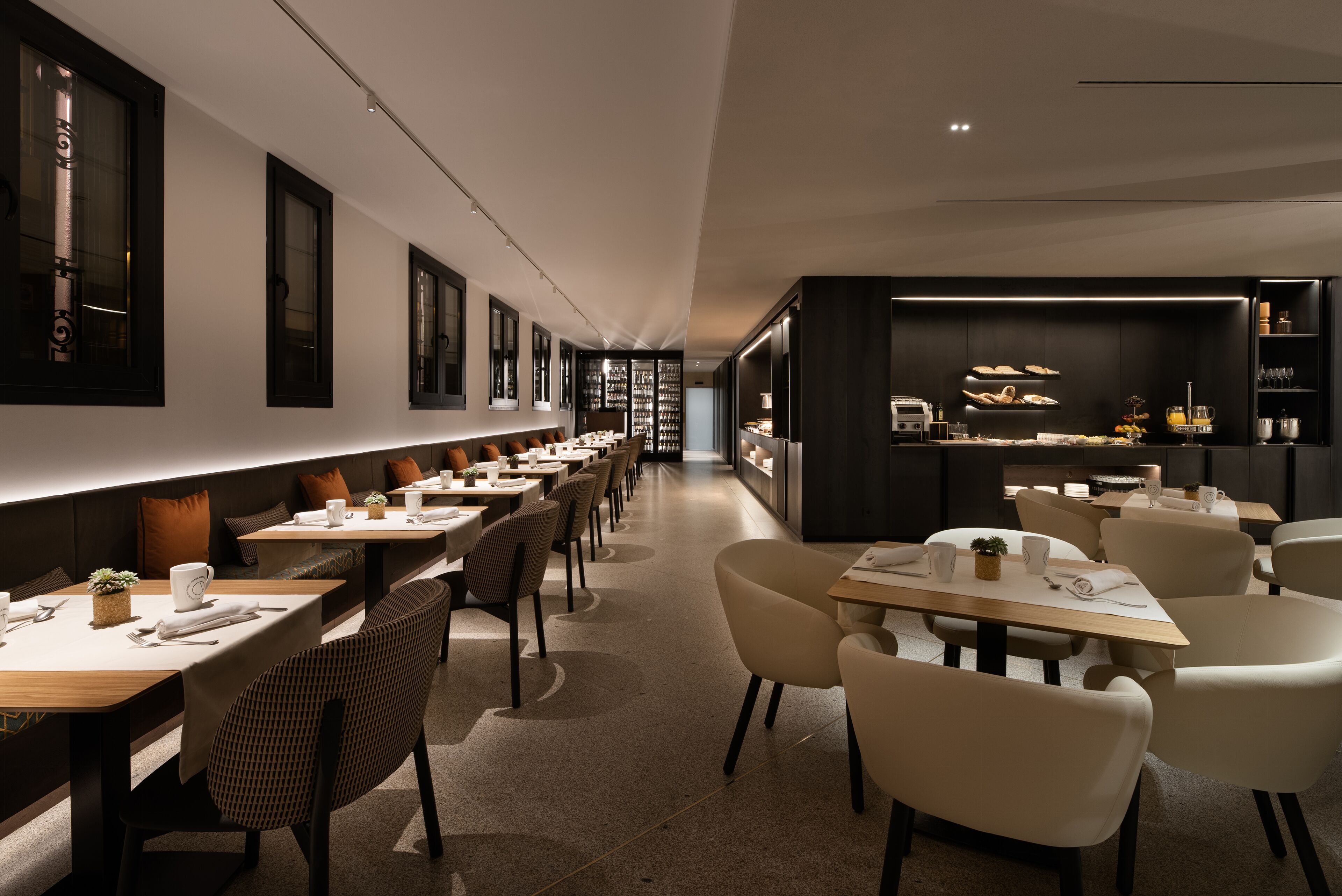 Un restaurant moderne avec de longues tables et des chaises, un espace cuisine et un long comptoir où sont exposés divers plats.