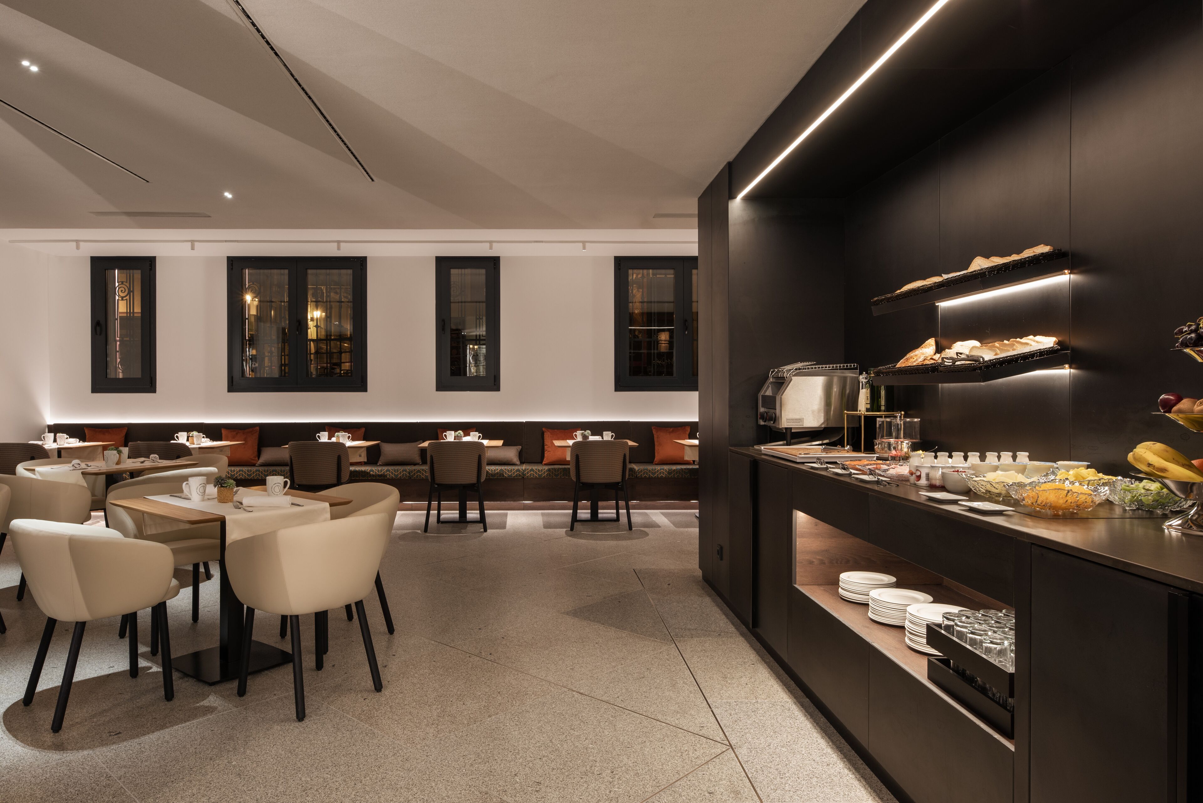Un espace repas moderne avec des tables et des chaises rondes, un buffet proposant divers plats, et un coin salon avec des canapés et des coussins.