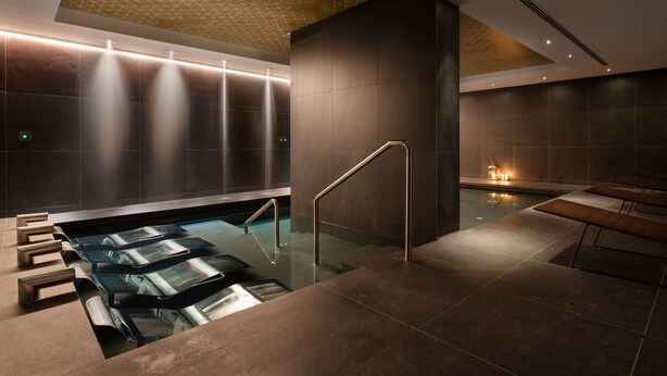 Ein moderner Wellnessbereich mit einem gläsernen Whirlpool, einer Lounge und einem geometrischen Deckenmuster.