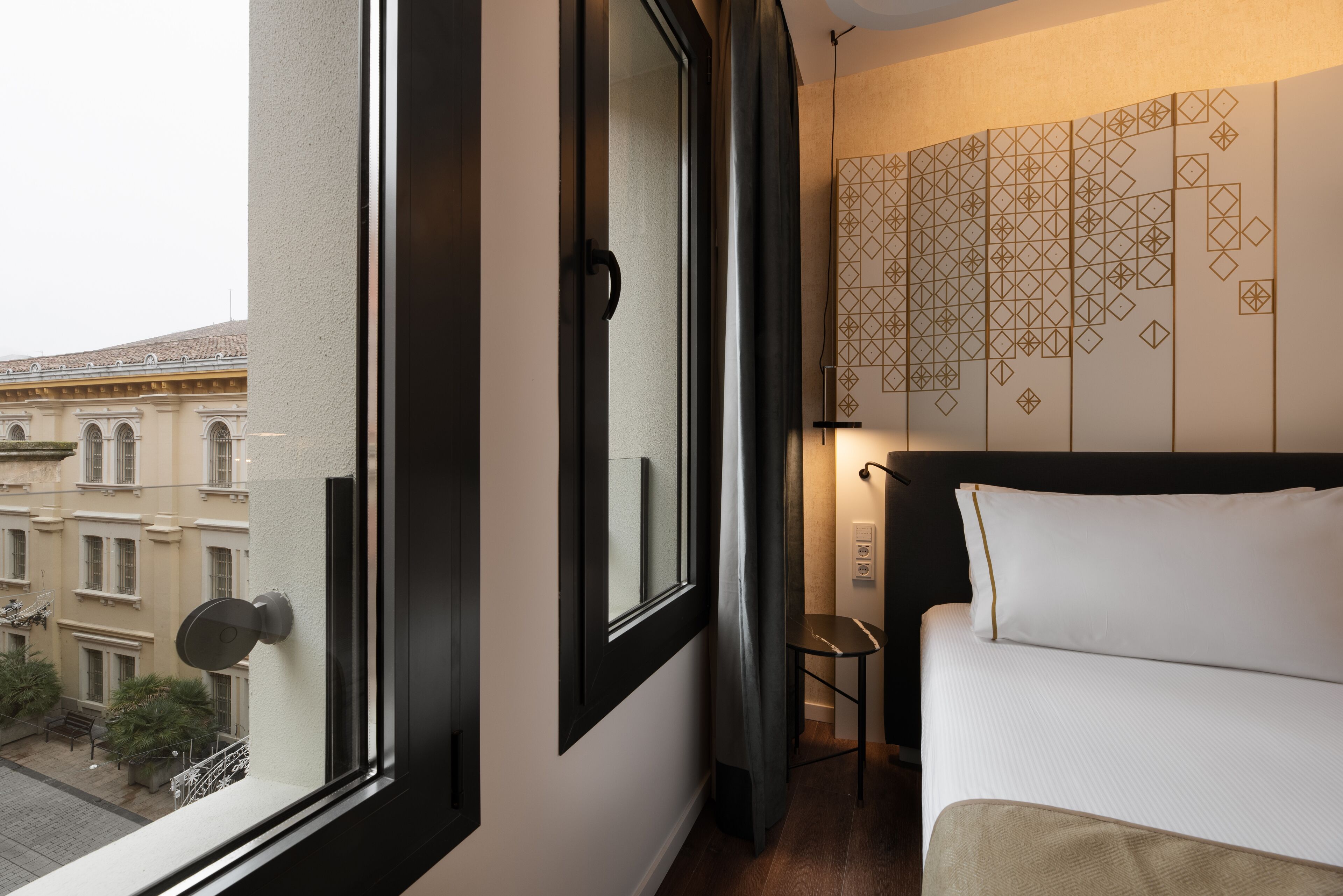 Une chambre d’hôtel avec une grande fenêtre, un lit recouvert de draps blancs, une petite table et un mur orné de motifs géométriques.