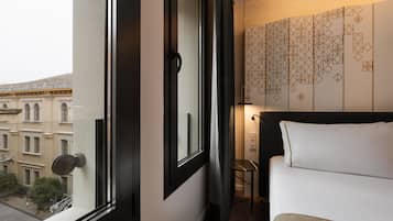 Une chambre d’hôtel avec une grande fenêtre, un lit recouvert de draps blancs, une petite table et un mur orné de motifs géométriques.