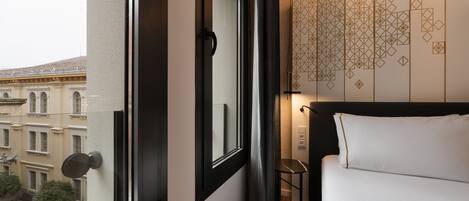 Habitación de hotel con un ventanal grande, una cama con ropa de cama blanca, una mesita y una pared con diseños geométricos.