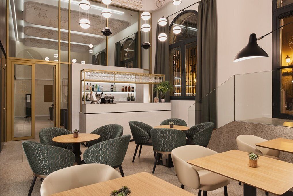 Un restaurante moderno con mesas redondas y sillas tapizadas de verde, una zona de bar con estanterías llenas de botellas y una escalera que lleva a un nivel superior.