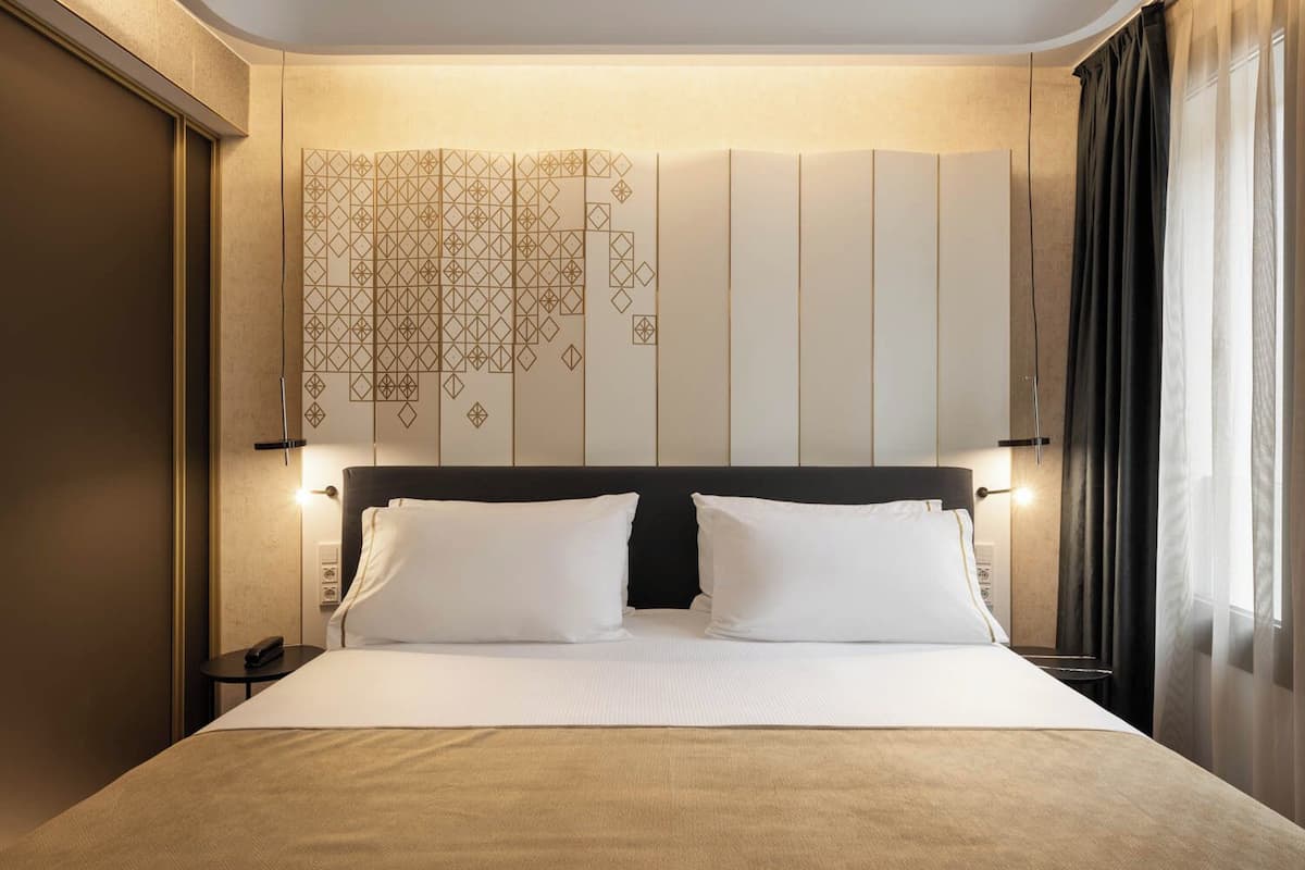 Ein Hotelzimmer mit einem großen Bett, einem Kopfteil mit geometrischen Mustern, Nachttischlampen und Blick ins Freie durch ein Fenster mit Vorhängen.