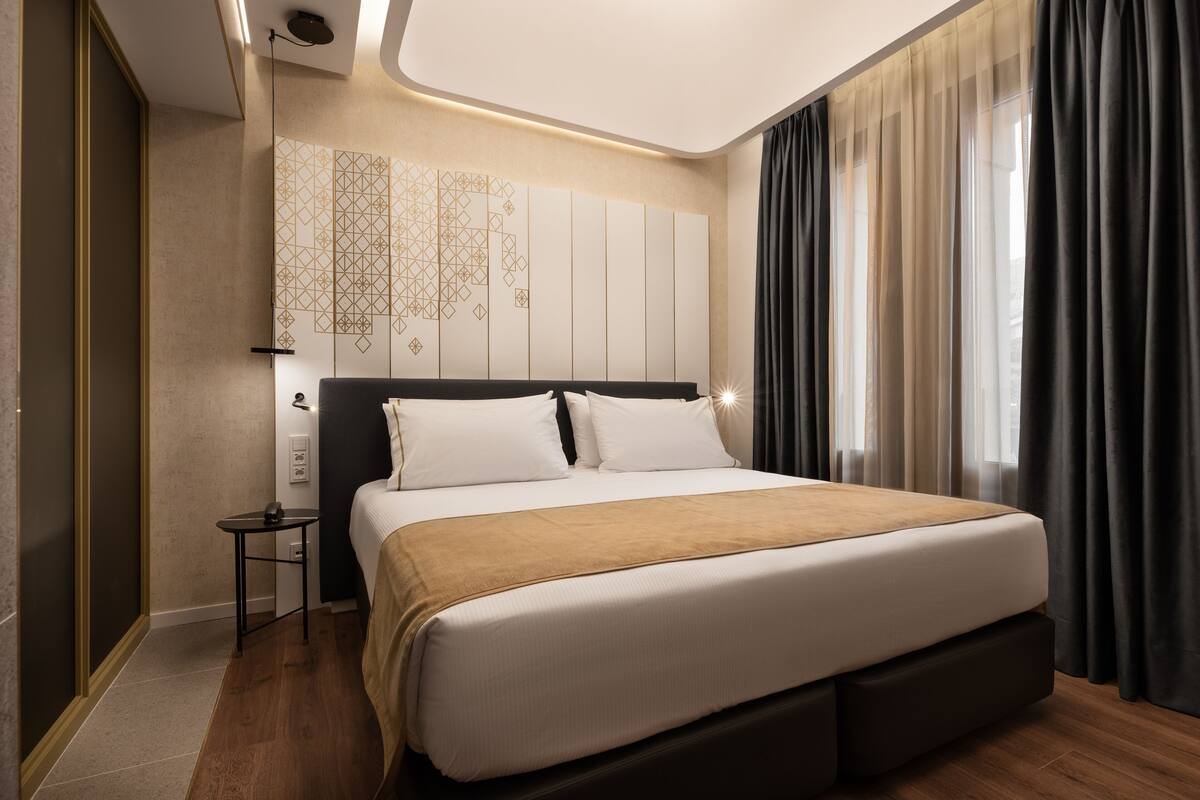 Ein modernes Hotelzimmer mit einem großen Bett, einem Nachttisch, einem an der Wand montierten Fernseher und einem Fenster mit Vorhängen.