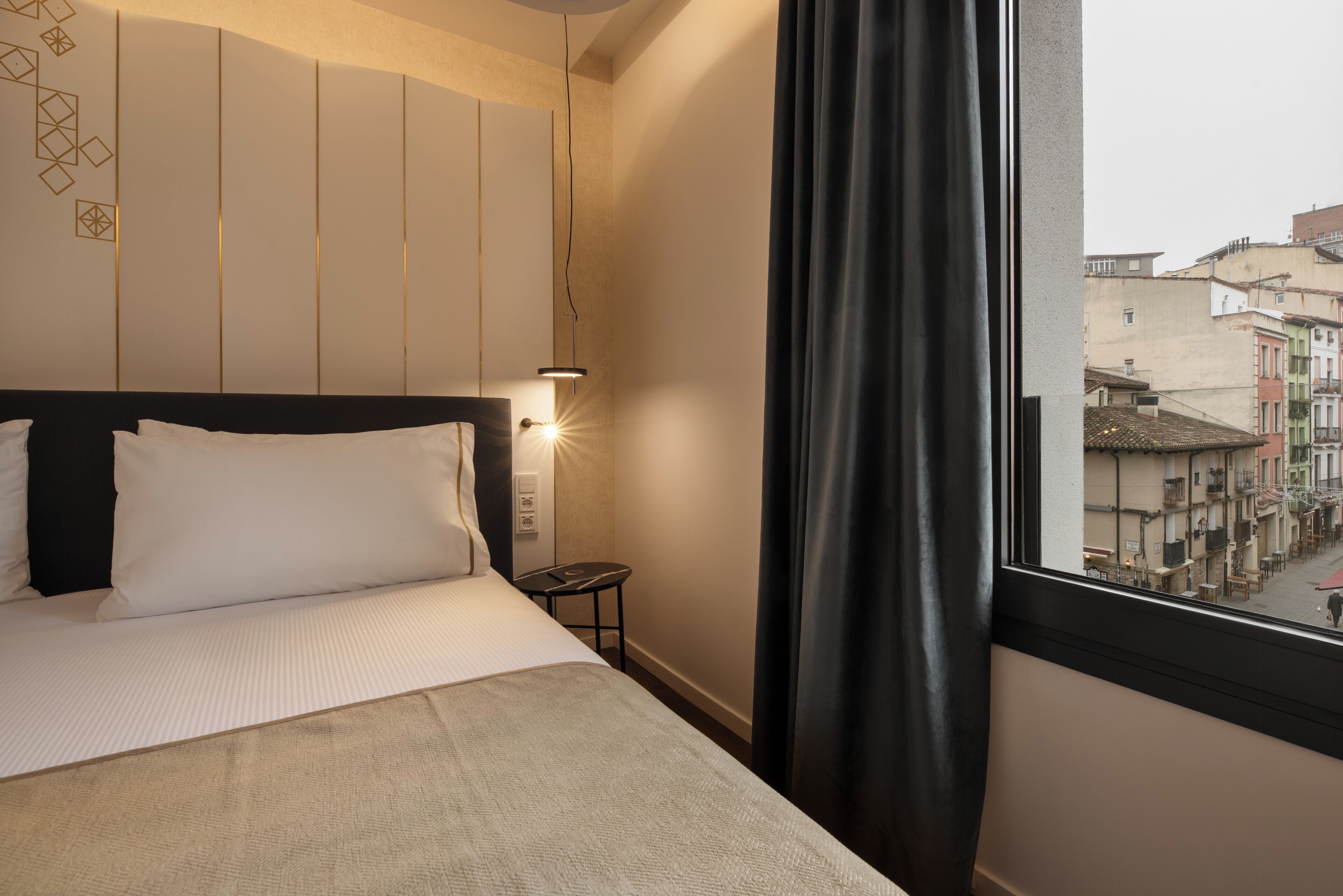 Una camera d'albergo con un letto, un comodino, una lampada e vista sulla città attraverso una finestra.