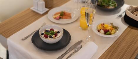 Un desayuno con un tazón de yogur cubierto de frutos rojos, un vaso de jugo de naranja y un plato de ensalada de frutas.