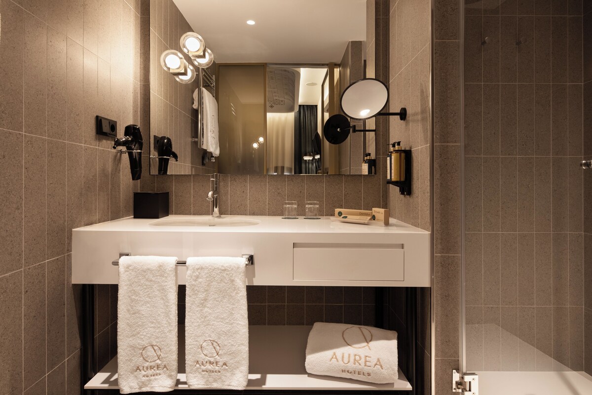 A modern bathroom with a large mirror, a sink, and two towels with "Aurea Hotels" branding.