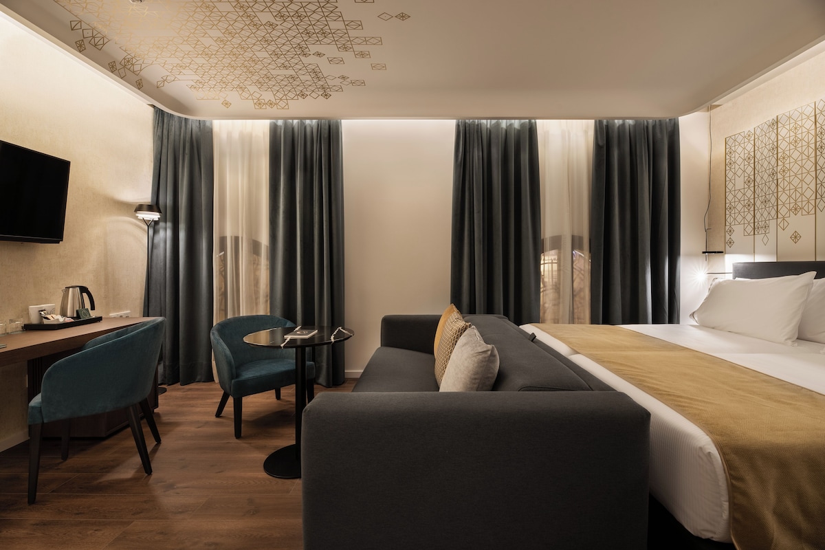 A modern hotel room with a bed, sofa, desk, and chairs.