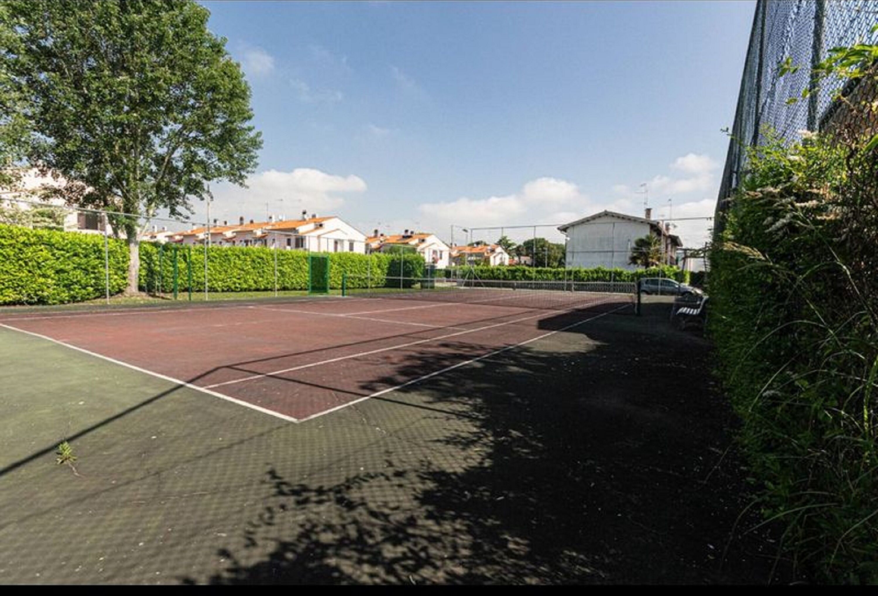 Un court de tennis en plein air, doté d’un filet, entouré de verdure et de bâtiments résidentiels.