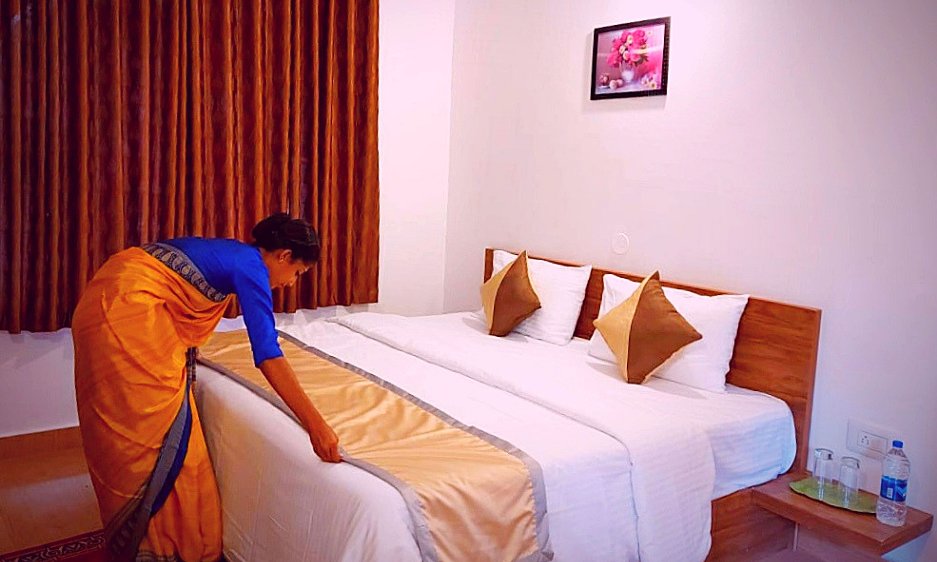 A hotel room with a bed, pillows, a nightstand with a water bottle, and a picture on the wall.