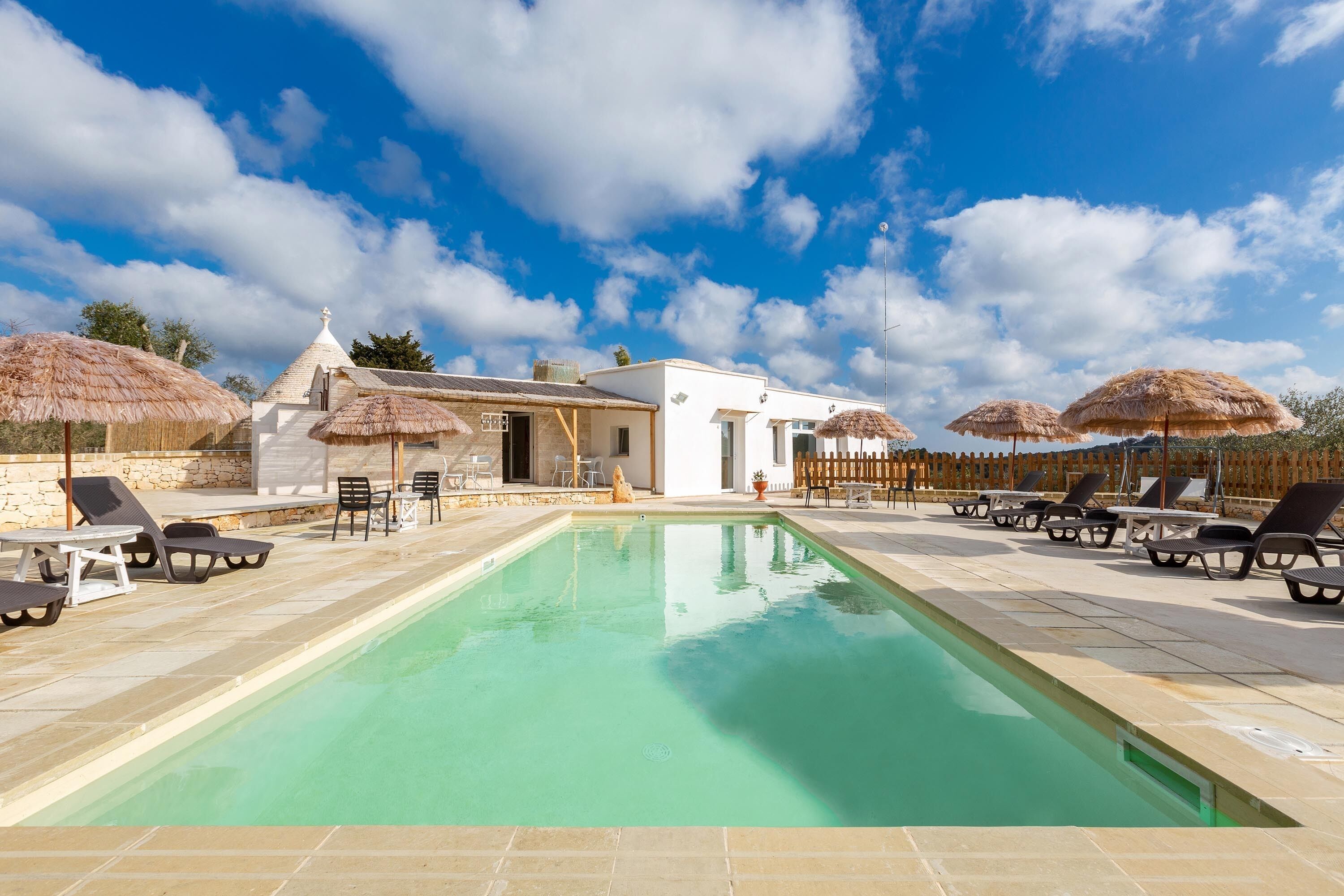 Área da piscina com espreguiçadeiras e guarda-sóis, um edifício com fachada branca e céu azul.