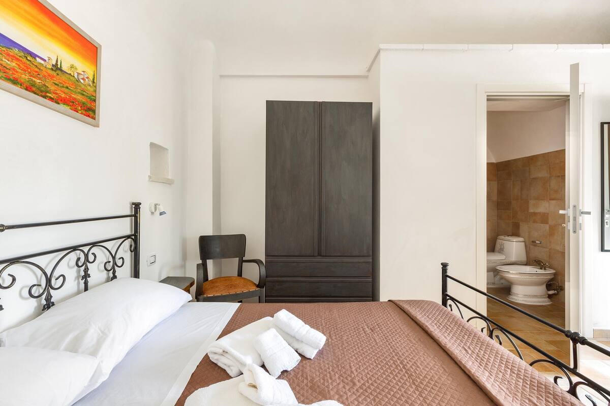 Une chambre d’hôtel avec un lit, une chaise, une armoire et une salle de bain visible à travers une porte ouverte.