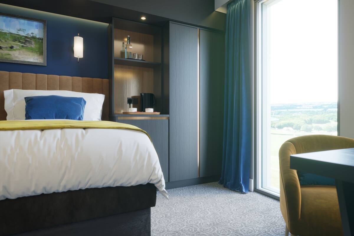 A hotel room with a bed, a desk, a chair, a window with a view, and a shelf with items.