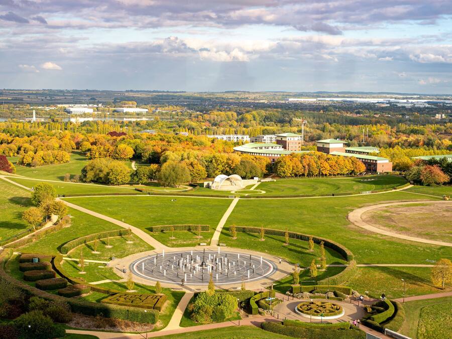 An aerial view of a park with a circular fountain, pathways, and surrounding greenery.