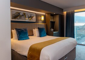 Una habitación de hotel moderna con una cama grande, cabecera con iluminación integrada, mesita de noche con una botella y vasos, y vista a la ciudad a través de la ventana.