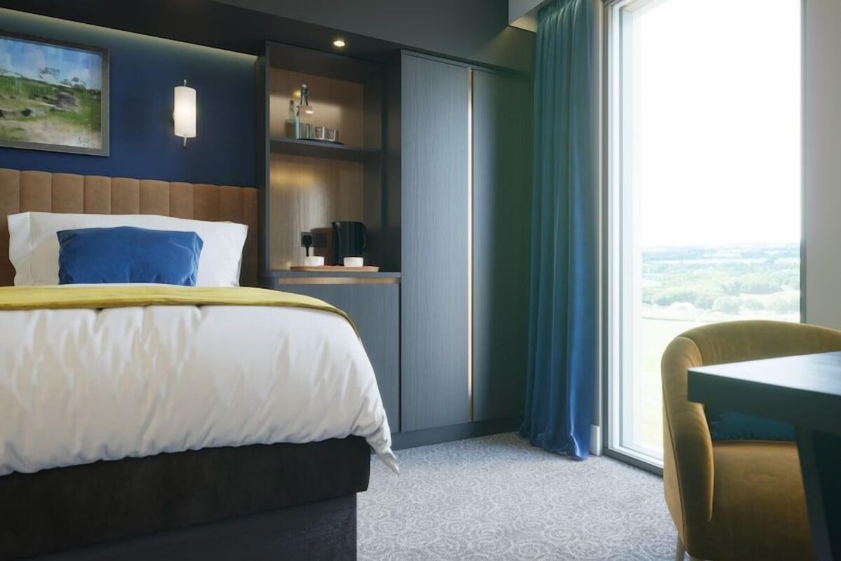 A hotel room with a bed, a desk, a chair, a window with a view, and a shelf with items.