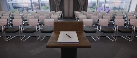 Un salón de conferencias con filas de sillas, una mesa de madera con un bloc de notas y un bolígrafo, y ventanas amplias con cortinas.