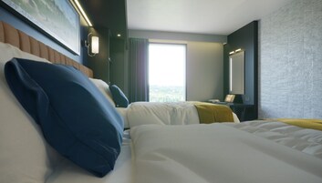 Habitación de hotel con dos camas, un ventanal grande, un cuadro en la pared y una mesita de noche con lámpara.