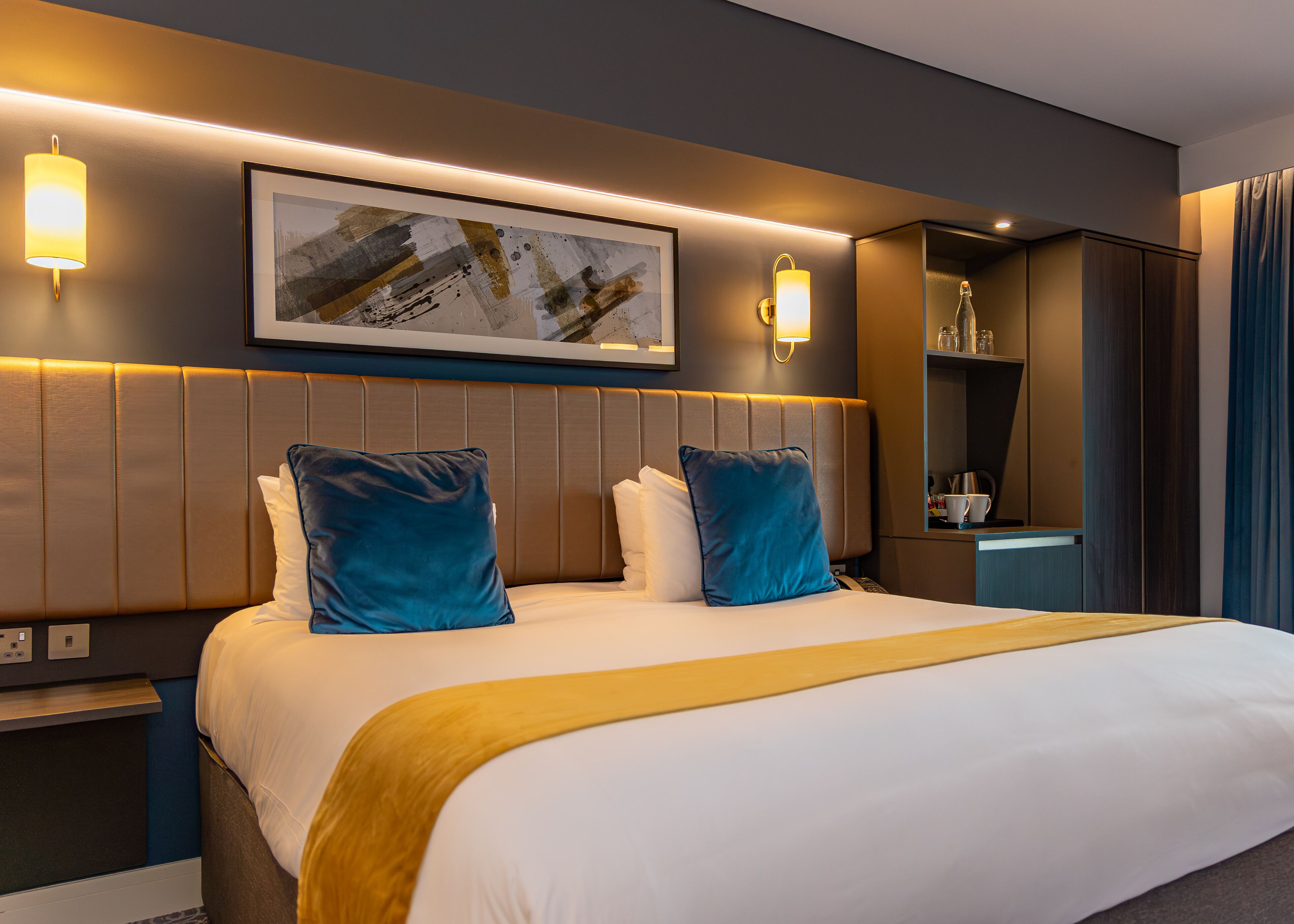 A modern hotel room with a large bed, two blue pillows, a gold throw blanket, and a framed abstract painting above the bed.
