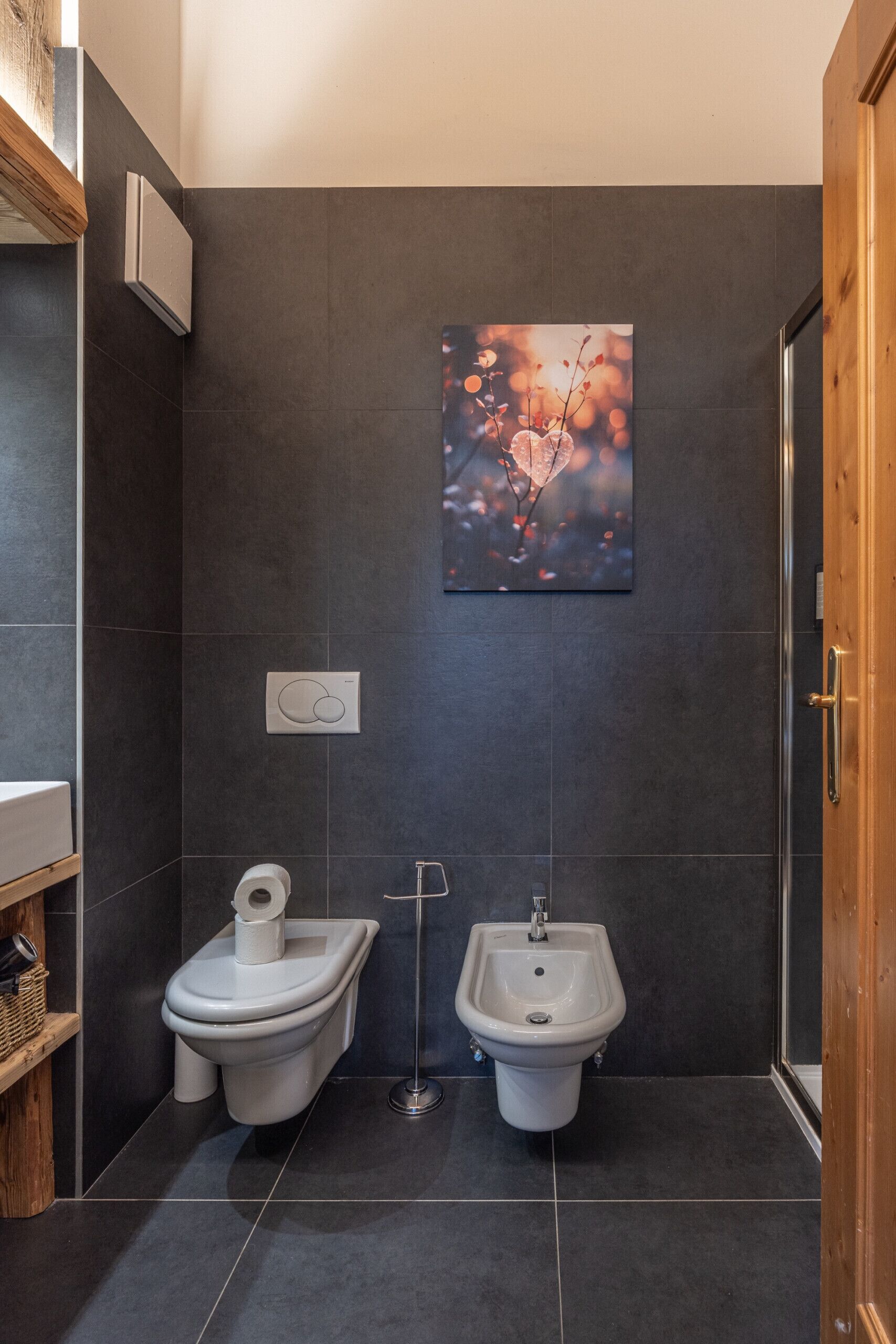 Un baño moderno con azulejos oscuros, un bidé, un inodoro, un ambientador de pared y un cuadro enmarcado.